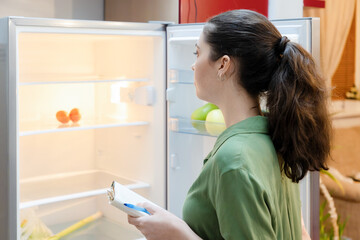 Back view of young Caucasian woman looks into empty refrigerator to make grocery shopping list. Housewife's chores