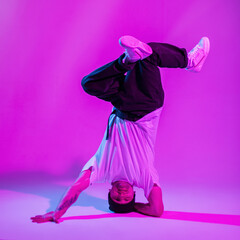 Cool fashionable professional male breakdancer in trendy fashion clothes with hat, t-shirt and sneakers stands on head and dances in a creative color studio with pink and neon lights
