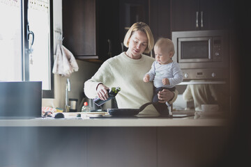 Fototapeta na wymiar Happy mother and little infant baby boy together making pancakes for breakfast in domestic kitchen. Family, lifestyle, domestic life, food, healthy eating and people concept