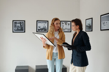 Two female workers of art gallery discussing restored picture and deciding where to hang it while preparing for opening of exhibition - obrazy, fototapety, plakaty