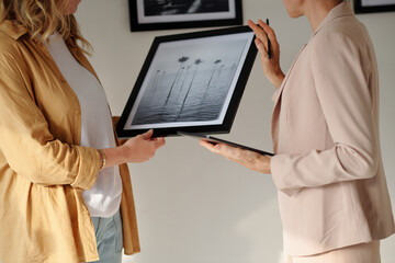 Close-up of two female restorers discussing black-and-white artwork in frame while choosing new...