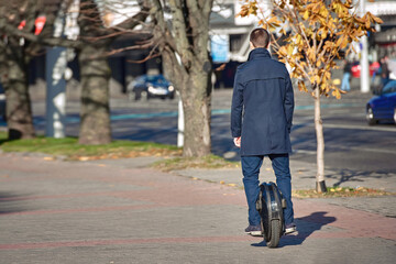 Trendy man in coat on electric monowheel riding fast (EUC). Man riding electric unicycle on city street, commute to work on electric transport. Commuter rides motorised unicycle along city street