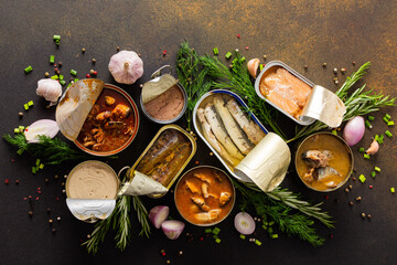 Different open tin cans with canned fish among spices and herbs on a brown background, canned...