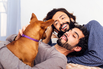 gay male couple smiling and relaxing happily lying in bed together with their little dog
