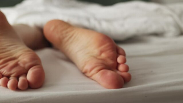 Person's legs sticking out from under blanket. Female barefoot close-up with dry calluses and corns. Zoom out defocused shot.