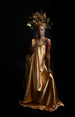  fantasy portrait of beautiful african woman model with afro, goddess silk robes and ornate floral wreath crown. gestural Posing holding golden flowers. isolated on dark  studio background 