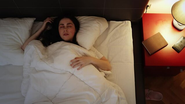 Insomnia and stress. Young Caucasian sleppy woman tosses and turns in bed. Top view. Electric evening light.