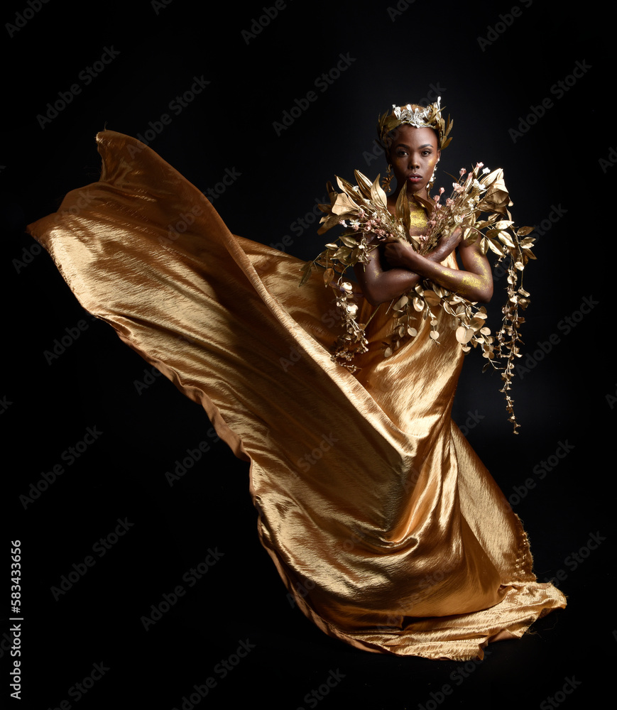 Wall mural  fantasy portrait of beautiful african woman model with afro, goddess silk robes and ornate floral wreath crown. gestural Posing holding golden flowers. isolated on dark  studio background 