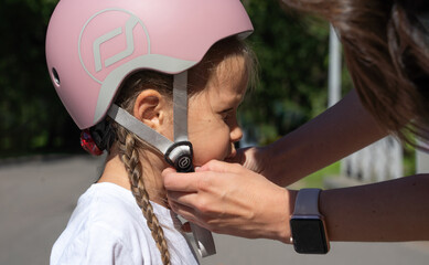Mom putting on inline skates rollers protection helmet to daughter child in public park in summer....