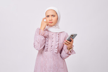 Pensive beautiful young Asian Muslim woman wearing hijab and purple dress holding mobile phone and...