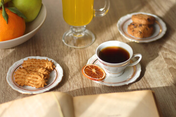 Cup of tea, plates with cookies, glass of orange juice, books, reading glasses, bowl of fruit and candles on the table. Selective focus.