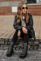Fashionable  blonde woman model with  black leather jacket and style sunglasses sitting on a floor at the city 