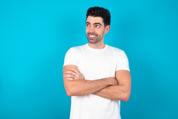 Fototapeta premium Dreamy rest relaxed Young man wearing white T-shirt over blue studio background crossing arms, looks good copyspace