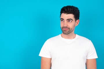 Young man wearing white T-shirt over blue studio background looking aside into empty space thoughtful