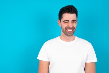 Coquettish Young man wearing white T-shirt over blue studio background smiling happily, blinking at camera in a playful manner, flirting with you.