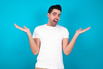 Naklejka premium Young man wearing white T-shirt over blue studio background celebrating crazy and amazed for success with arms raised and open eyes screaming excited. Winner concept