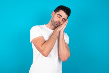 Fototapeta na wymiar Young man wearing white T-shirt over blue studio background sleeping tired dreaming and posing with hands together while smiling with closed eyes.