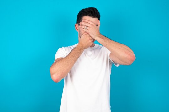 Young man wearing white T-shirt over blue studio background Covering eyes and mouth with hands, surprised and shocked. Hiding emotions.