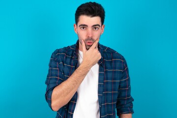 Young man standing over blue studio background Looking fascinated with disbelief, surprise and amazed expression with hands on chin