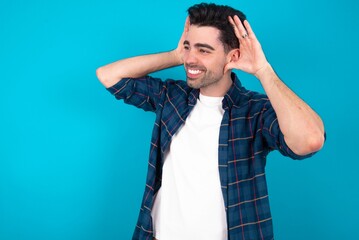 Young man standing over blue studio background Trying to hear both hands on ear gesture, curious for gossip. Hearing problem, deaf