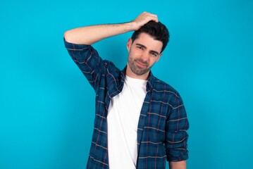 Young man standing over blue studio background being confused and wonders about something. Holding hand on head, uncertain with doubt. Pensive concept.