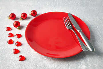 Empty red ceramic plate with cutlery surrounded by red Christmas balls and hearts on a light...
