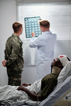 Professional doctor pointing at x-ray image and discussing it with soldier with wounded man lying on bed in ward