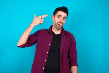 Young man standing over blue studio background foolishness around shoots in temple with fingers makes suicide gesture. Funny model makes finger gun pistol