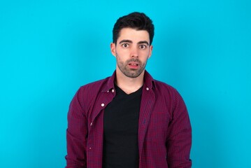 Obraz premium Shocked Young man standing over blue studio background stares bugged eyes keeps mouth opened has surprised expression. Omg concept