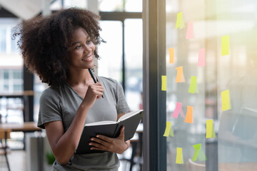 Young smiley attractive, businesswoman using sticky notes on a glass wall to write a strategic business plan to develop and grow to success.