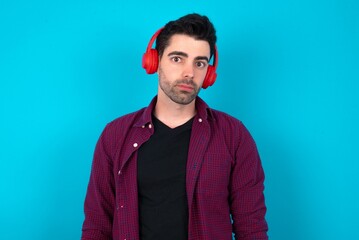 Serious displeased Young man standing over blue studio background looks puzzled at camera being angry wears stereo headphones listens music while walking at street