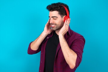 Young man standing over blue studio background with headphones on her head, listens to music, enjoying favourite song with closed eyes, holding hands on headset.