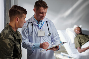 Doctor pointing in medical card and discussing diagnosis of patient together with soldier in...