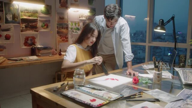 Zoom in shot of young Asian woman painting picture on paper with help of male teacher while taking lesson in art school in evening