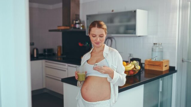 A young pregnant woman drinks a prepared vitamin smoothie and uses a smartphone standing in the kitchen at home. Healthy eating during pregnancy.