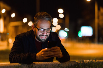 A happy man with glasses is using a smartphone in the city with a nightlight in the background.