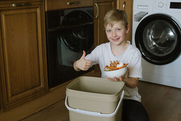 Happy Smiled Blond Boy In Kitchen Making Compost Scraping Vegetable Leftovers Into Bin at Home. Zero waste concept. Processing and recycling organic.