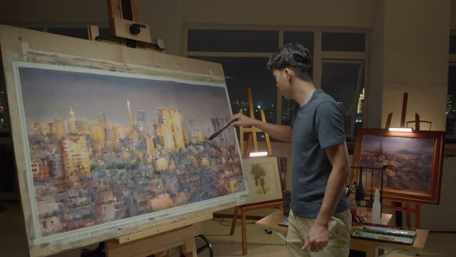 Medium long shot of young Asian painter standing by easel and drawing panorama of night city on canvas while working late in creative studio