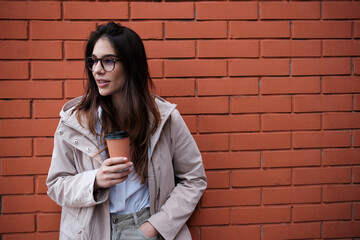 A happy smart casual woman leaning on brick wall outside and enjoying takeaway coffee.