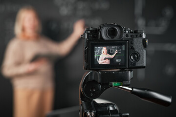 Mature elegant woman pointing at blackboard and recording lesson on professional camera