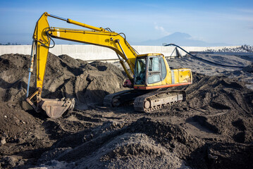 A yellow big excavator stands on a construction site with dark soil on a sunny day with a blue sky.