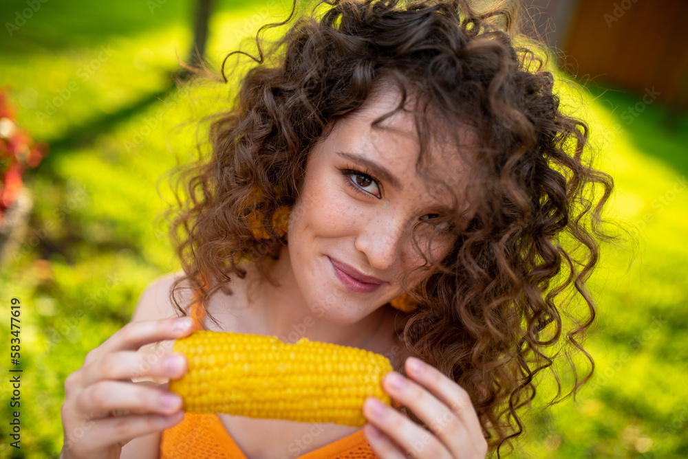 Wall mural a beautiful girl eats delicious corn against a background of green grass. curly girl eats corn.