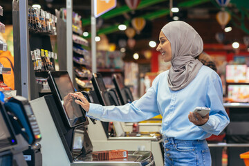 A woman buyer in a supermarket in a hijab pays for goods at a self-service checkout, convenient...