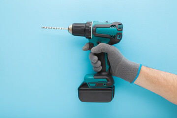 Young adult man hand holding and showing dark black green professional battery driver drill on light blue wall background. Pastel color. Closeup. Tool for repair work. Side view.