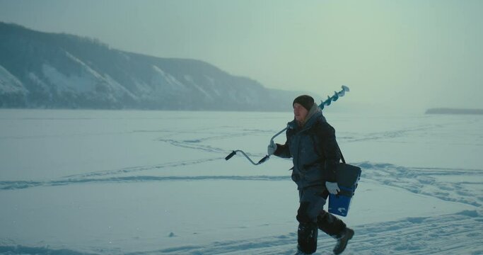 tired fisher with ice drill and tackle box walking on snowed frozen river in winter, 4K, Prores