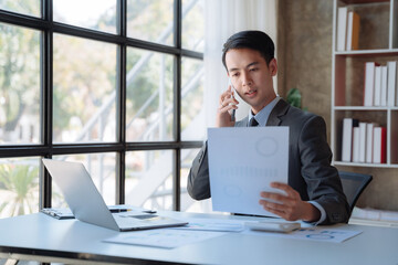 young Asian businessman involved in cellphone call conversation working on computer, giving professional consultation to client or negotiating project.