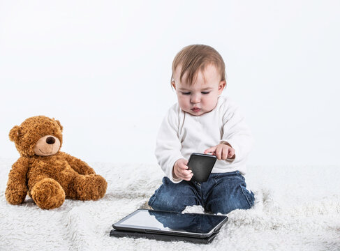 Studio photo with a white background of a baby touching the screen of a tablet with a teddy bear on the side