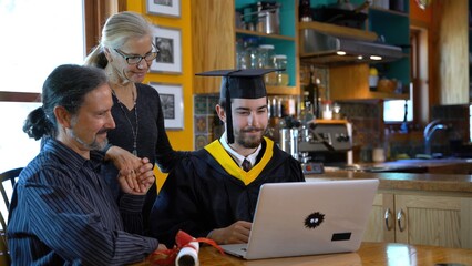 Family watches computer as son, dressed in graduation costume switches sides with tassel and then picks up diploma and smiles. Everyone hugs.