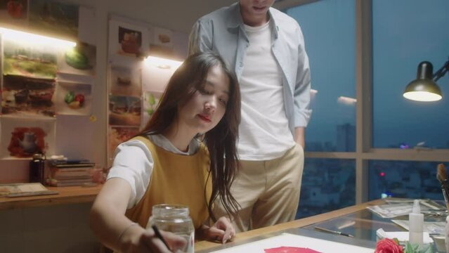 Medium shot of Asian male artist teaching female student as she painting picture on paper during class in art school in evening