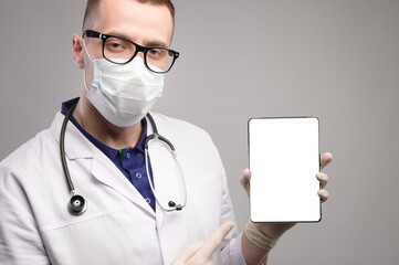young and handsome smart doctor wearing a surgical hygiene protective mask on a gray background and holding a tablet computer in his hand with a blank mockup screen. Idea for doctors.